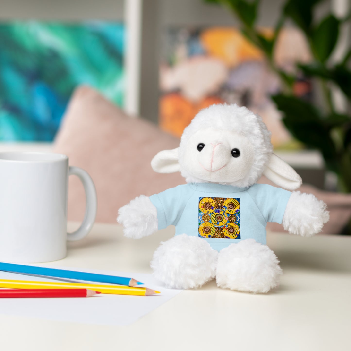 White plush lamb toy wearing a blue shirt with a colorful design, sitting on a table with a mug and pencils.