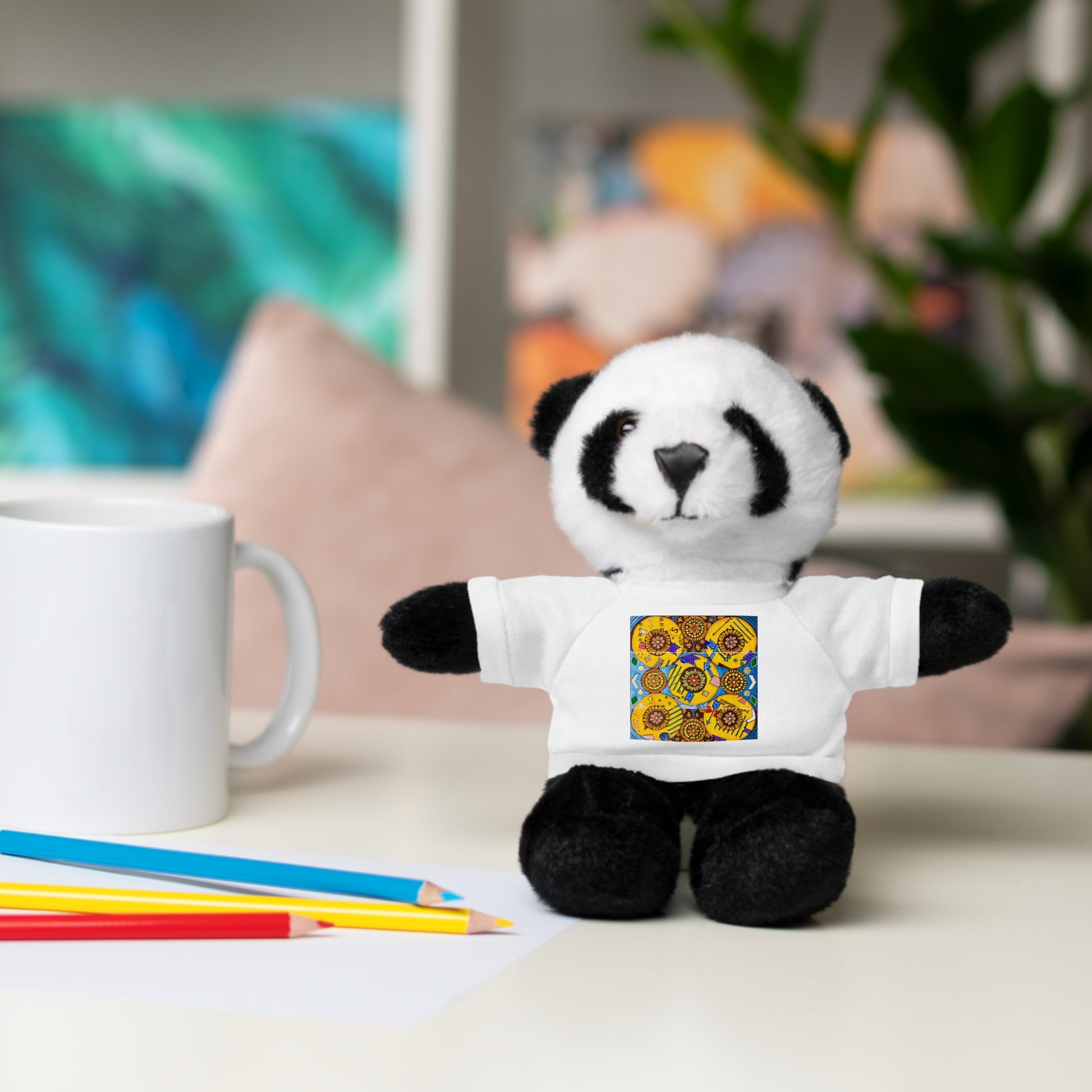 Plush panda wearing a white t-shirt with a colorful design, sitting on a table with stationery items.